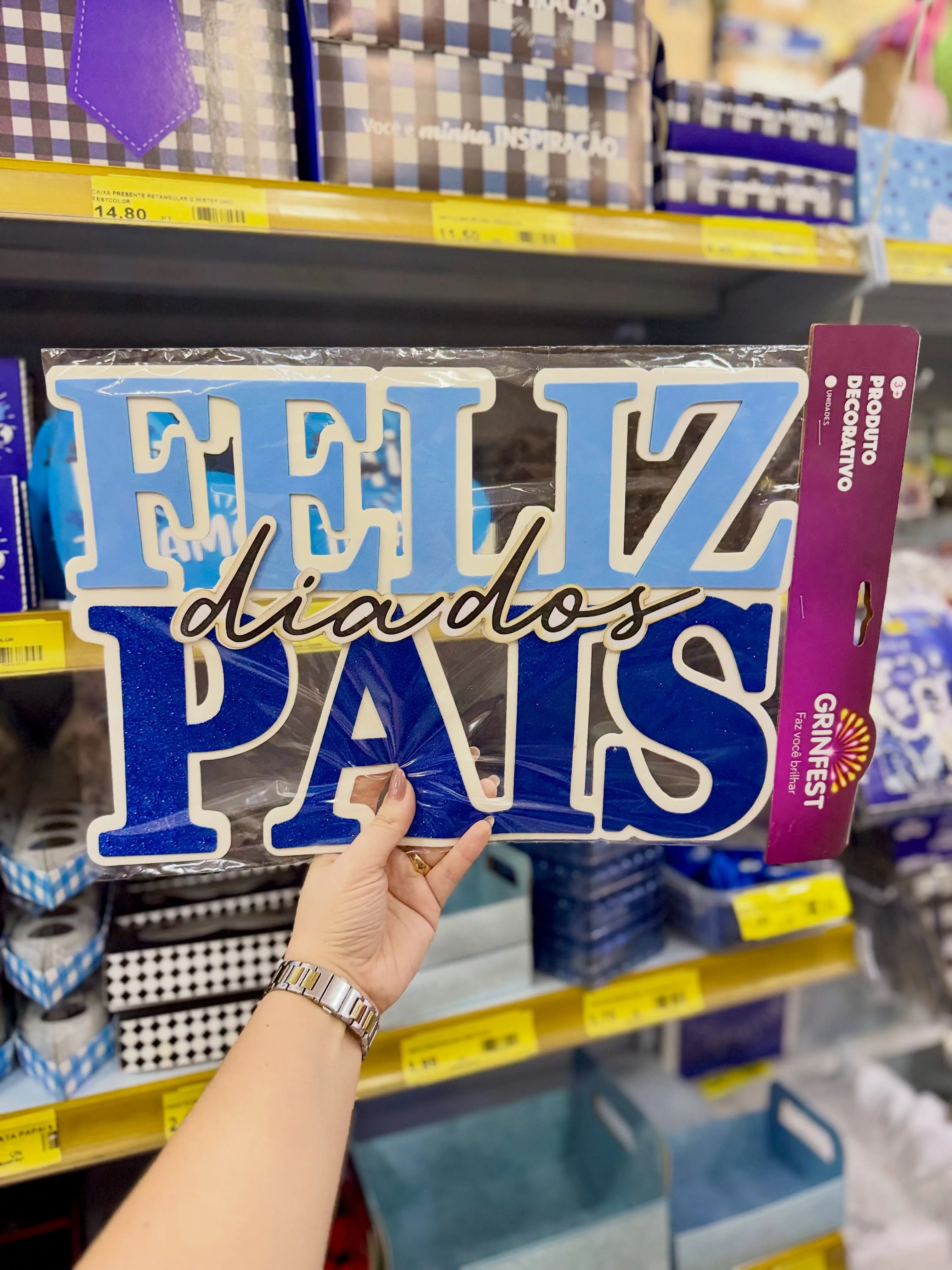 Mão segurando uma decoração de Dia dos Pais diante do setor sazonal da Karol Festa